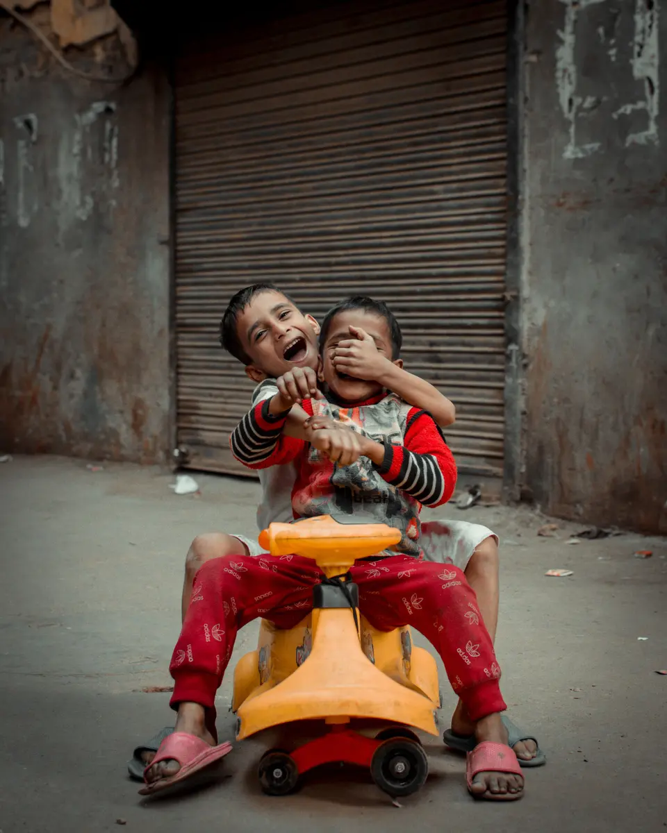 Two young Indian brothers laugh together while riding a shared plastic toy car on a street in New Delhi.