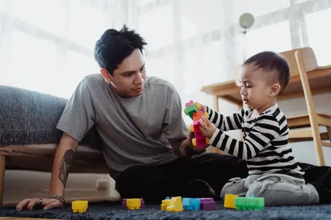A father sits on the floor at home with his toddler son, both hands-on together building a toy out of colorful plastic blocks.