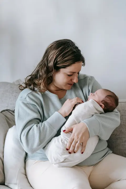 A mother sits upright on a sofa, cradling her sleeping newborn snugly against her chest.