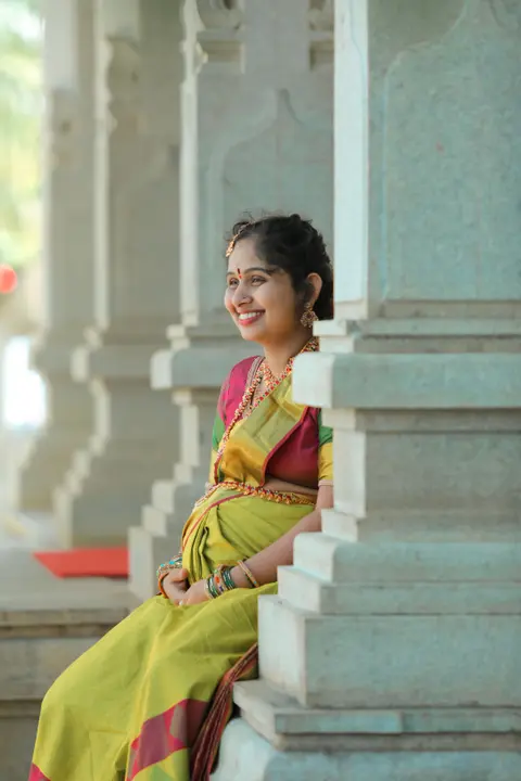 A pregnant South Asian woman in a colourful traditional saree sits smiling among bright stone temple columns, warm daylight.