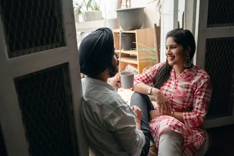 An Indian couple sits together on a doorstep at home, the husband holding a mug of coffee while both chat and smile.
