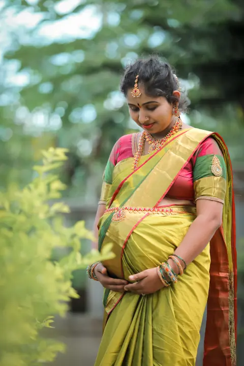 A pregnant woman in a yellow-green sari with gold jewelry looks down tenderly at her belly in a garden