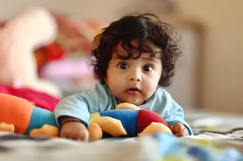 An Indian baby with curly dark hair lies on her stomach on a bed, awake and alert, reaching toward a blurred pink plush toy, wearing a light blue top.