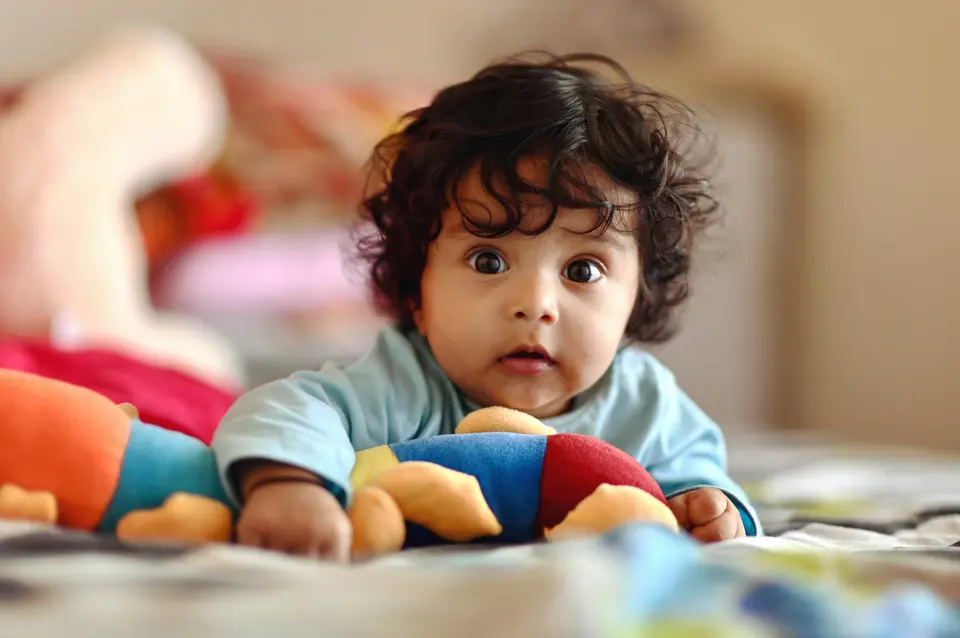 An Indian baby with curly dark hair lies on her stomach on a bed, awake and alert, reaching toward a blurred pink plush toy, wearing a light blue top.