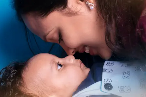 A South Asian mother leans close to touch noses with her smiling baby, who she holds upright and supported against her chest.