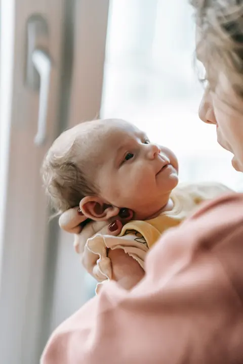 A mother holds her newborn close by a window, both gazing at each other, in a calm, uncluttered room with soft natural light.