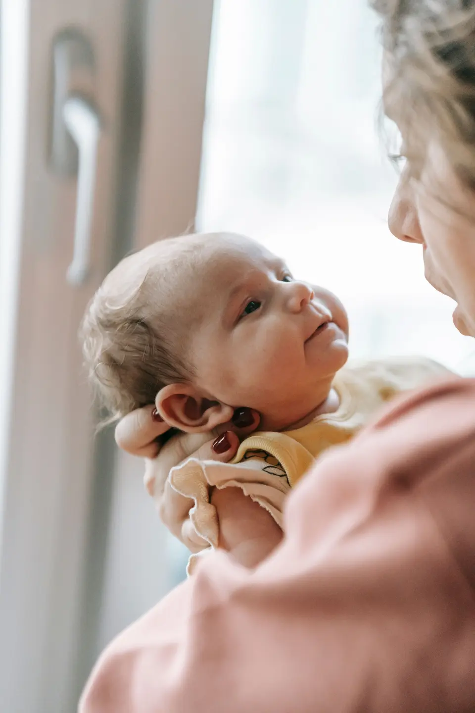 A mother holds her newborn close by a window, both gazing at each other, in a calm, uncluttered room with soft natural light.