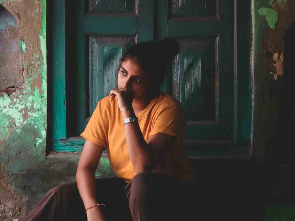 a young Indian woman sits in a thoughtful, tense pose with her hand near her chin, looking inward