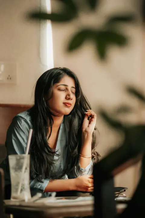 a young Indian woman sits with eyes closed, resting her head, looking fatigued in a cafe setting