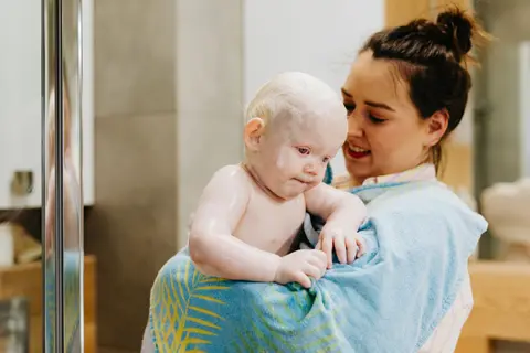 A mother smiles down at her toddler, freshly bathed and wrapped in a towel, in a bright bathroom doorway.