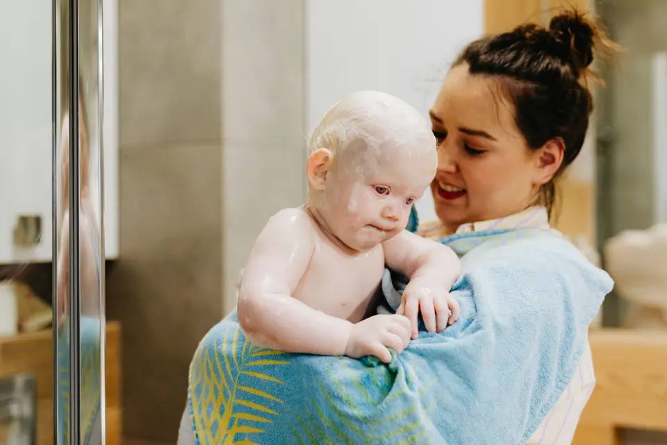 A mother smiles down at her toddler, freshly bathed and wrapped in a towel, in a bright bathroom doorway.