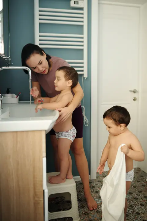 A toddler boy stands independently on a step-stool at a bathroom sink, his mother close beside him, while his younger brother watches nearby.