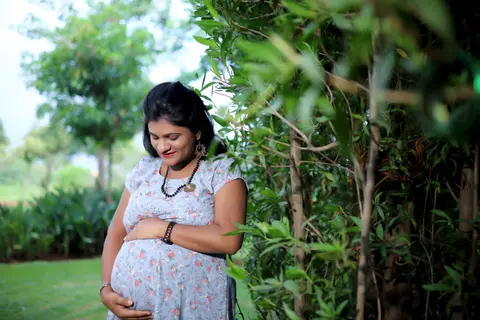 A pregnant South Asian woman in a simple floral day dress rests a hand on her belly, standing in a garden in soft daylight, no festive jewellery.