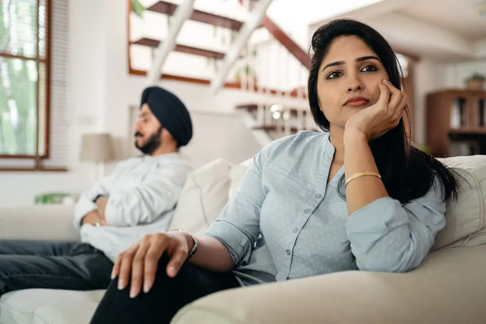 An Indian woman in a light blue shirt sits on a couch looking downcast and withdrawn, with a man in a black turban seated apart from her in the background