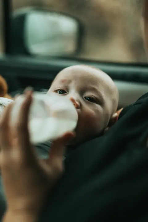 a baby held upright in a parent's arms drinks from an unbranded bottle of pumped milk