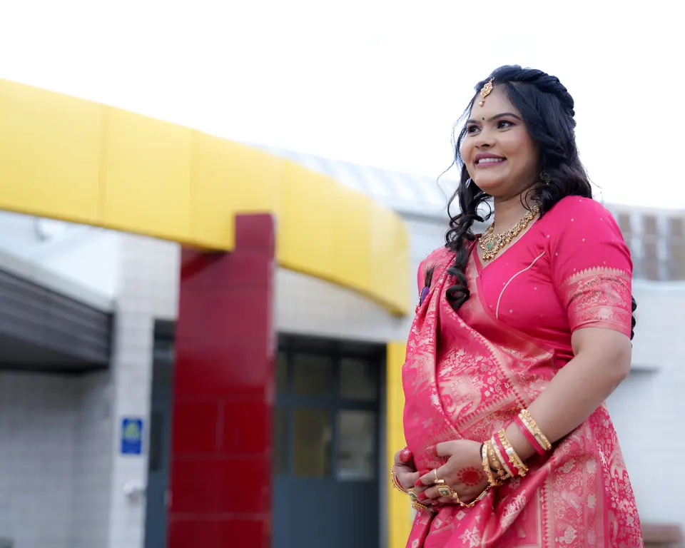 A smiling pregnant woman in a pink sari with gold jewelry stands outside a building, looking off to the side