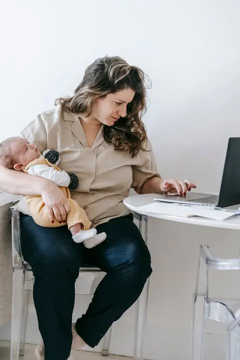 A mother works at a laptop on a small home desk while holding her sleepy newborn against her shoulder, plain white background.
