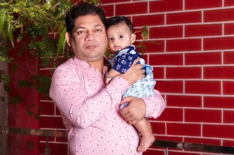 An Indian father holds his awake baby close against his chest outdoors, standing in front of a red brick wall with green leaves nearby.