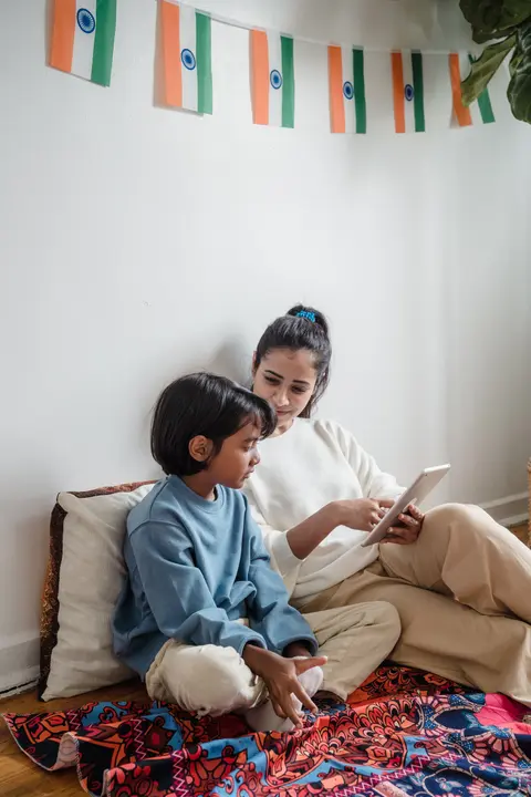 A South Asian mother and her toddler son sit cross-legged on a colorful rug at home, both looking at a tablet, with an Indian flag bunting on the wall.