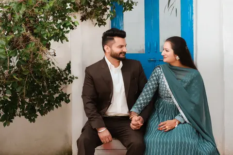A South Asian couple sit close together outdoors by a blue doorway, holding hands and smiling at each other, dressed in elegant semi-formal attire.