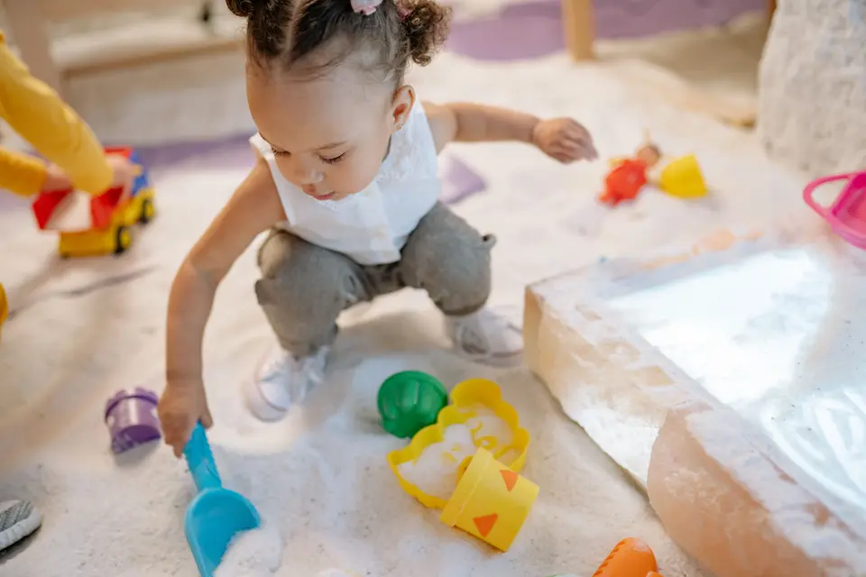 A toddler crouches to explore colorful toy molds and white play-sand with her hands in an indoor sensory play area.