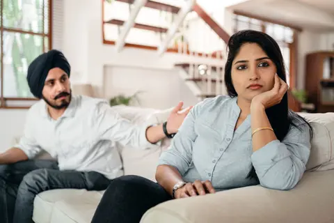 An Indian woman rests her cheek on her hand looking upset on a couch, while a man in a turban gestures beside her during a tense conversation