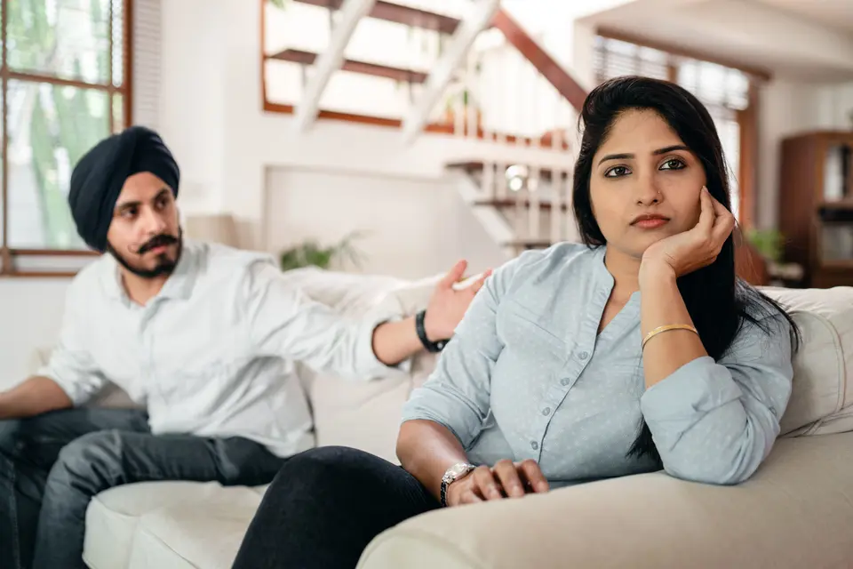 An Indian woman rests her cheek on her hand looking upset on a couch, while a man in a turban gestures beside her during a tense conversation