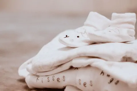 A folded stack of cream baby clothing printed with 'kisses' and 'hugs' script and a smiling bunny face, with a pair of baby socks resting on top.