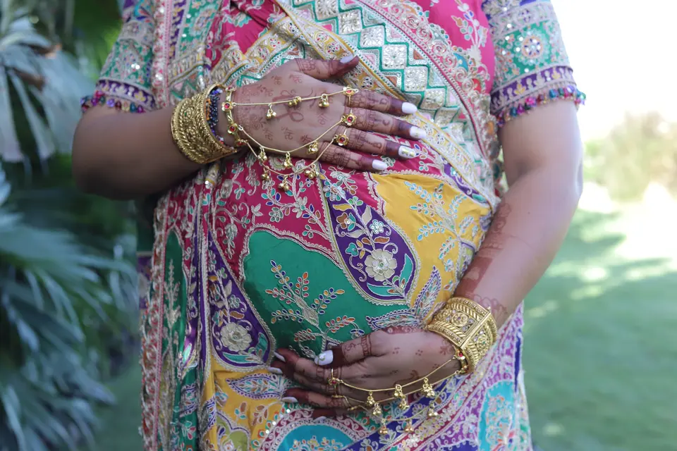 Close-up of a pregnant woman's henna-decorated hands resting on her belly, wearing a colorful embroidered sari and gold bangles