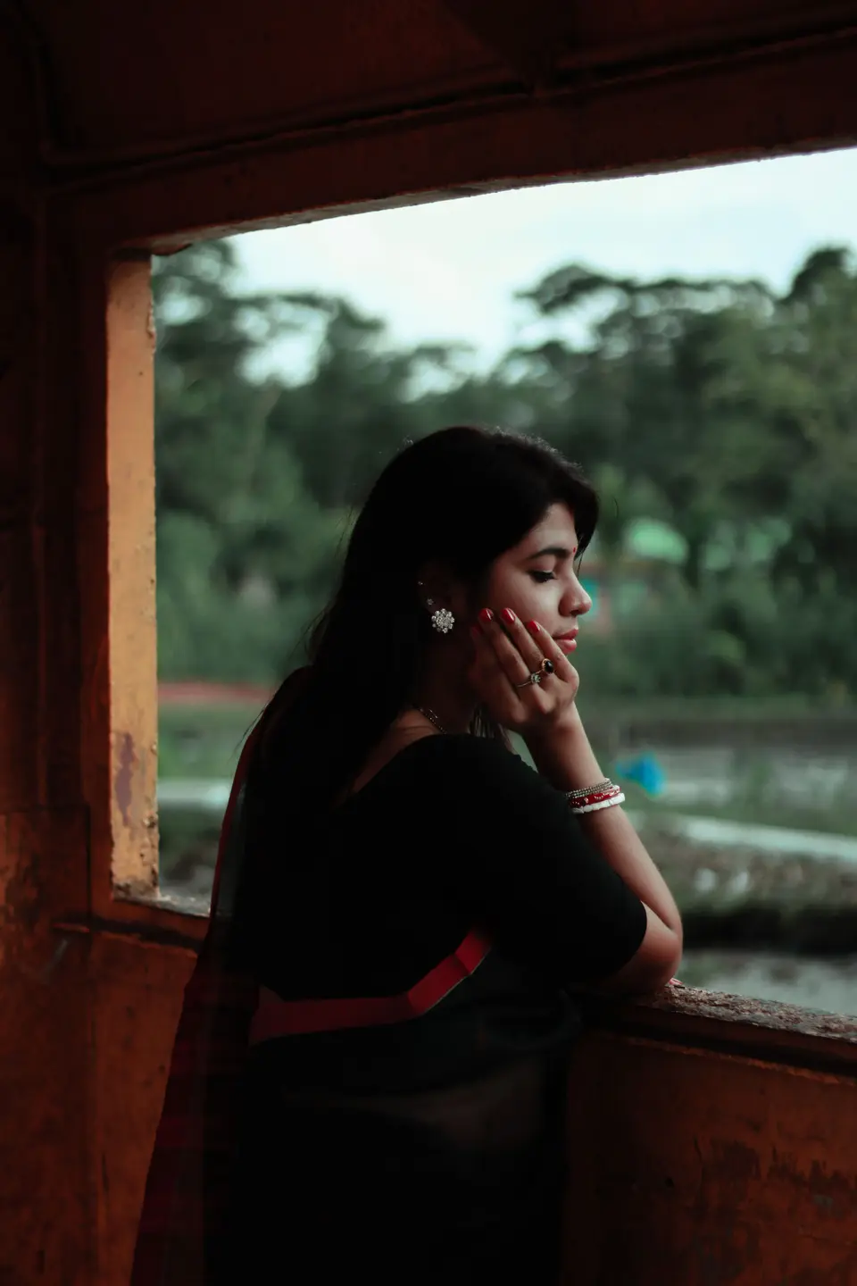 A young Indian woman in a black sari leans thoughtfully at a window, resting her hand near her chin.