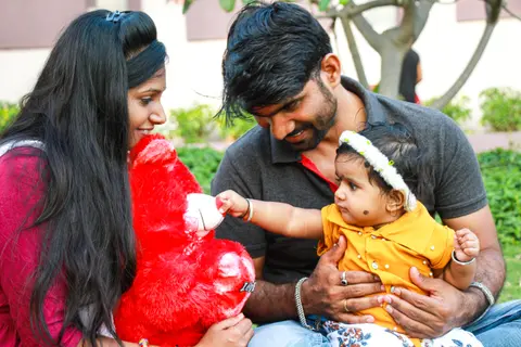 Indian parents sit together outdoors engaging their baby daughter, who reaches for a red stuffed toy her mother holds.
