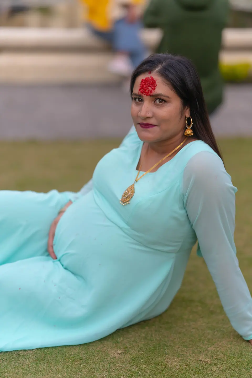 A pregnant woman in a turquoise dress lies back on grass, smiling gently with a red bindi and gold jewelry
