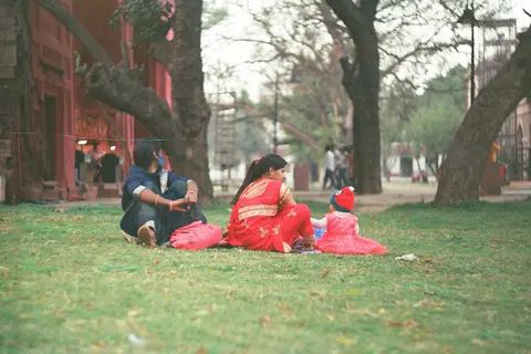two women in saris sit on the grass in a public park with a toddler, a simple, low-cost family outing