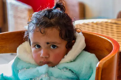 A South Asian one-year-old girl in a knit sweater sits in a wooden chair, hands resting on the armrests, looking at the camera.
