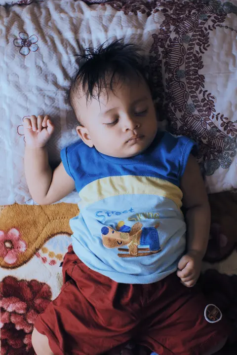 A South Asian baby naps on a floral bedspread, arms relaxed, wearing a blue-and-red outfit.