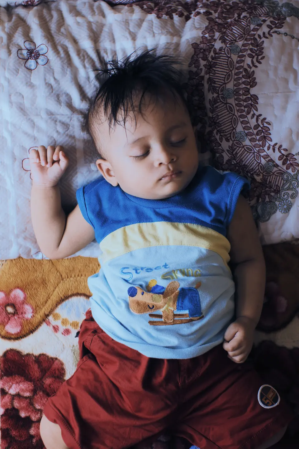 A South Asian baby naps on a floral bedspread, arms relaxed, wearing a blue-and-red outfit.