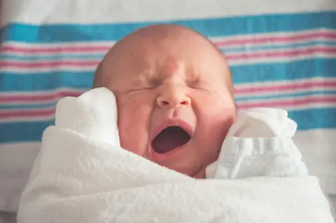 A newborn yawns widely with eyes scrunched shut, wrapped in a white towel against a blue-and-pink striped blanket.