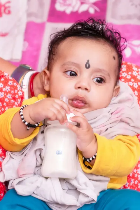 An Indian baby with a small forehead bindi drinks from a bottle held to his mouth by a caregiver, wrapped in a soft cream cloth.
