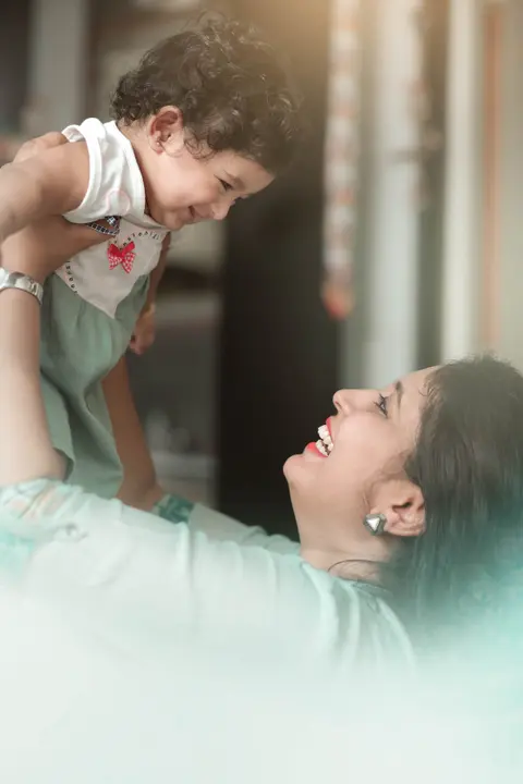 an Indian mother joyfully lifts her curly-haired baby up in the air indoors, both laughing, baby securely supported under the arms
