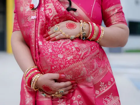 Close-up of a pregnant woman's hands with gold bangles resting on her belly in a bright pink embroidered sari