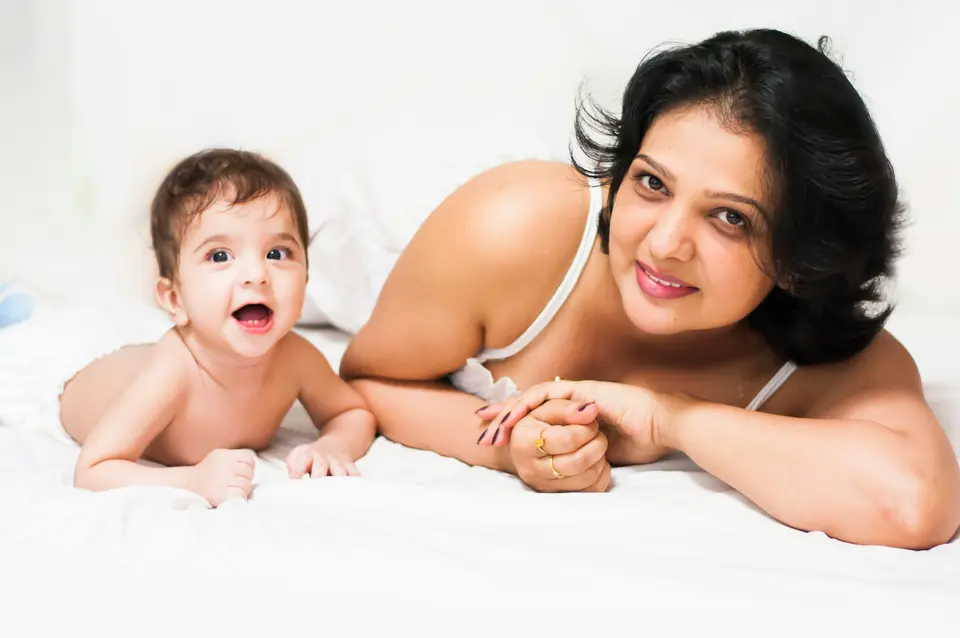 an Indian mother and her baby lie propped on their elbows on a white bed, both awake and smiling at the camera