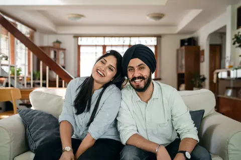 A laughing Indian couple sit close together on a couch, the woman leaning her head on the man's shoulder