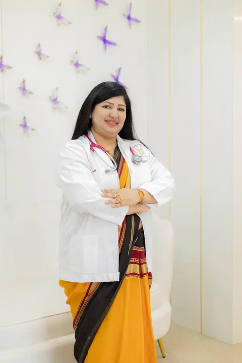 An Indian woman doctor in a white coat worn over a sari stands with arms crossed, a bindi and stethoscope visible.