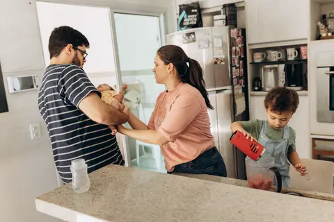 In a home kitchen, a father holds a newborn while a mother helps steady the baby's hand, as their older son pours a smoothie at the counter.