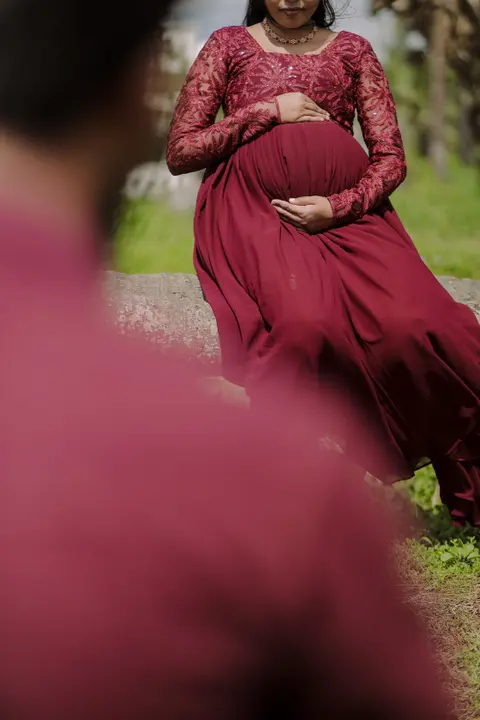 A pregnant woman in a maroon lace dress sits on a stone wall with her hands on her belly, a blurred figure in the foreground