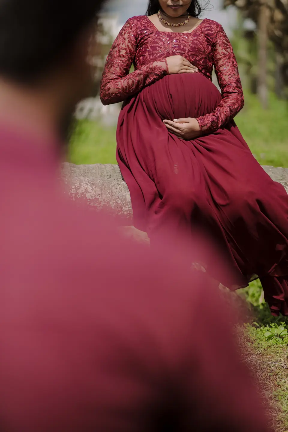 A pregnant woman in a maroon lace dress sits on a stone wall with her hands on her belly, a blurred figure in the foreground