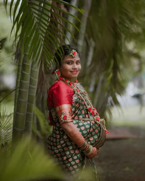 A smiling pregnant woman in a red-trimmed green sari with flowers in her hair stands among palm fronds, looking healthy and radiant