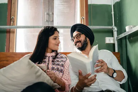 A Sikh couple sits together on a bed, the man holding a book, both smiling at each other.