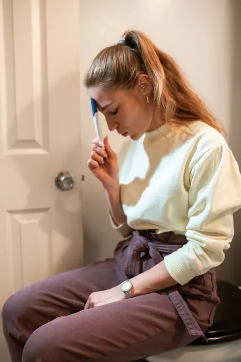 A woman sits indoors holding a pregnancy test to her forehead, eyes closed in thought.