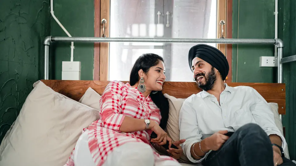 An Indian couple sits together on a bed, laughing and talking, one holding a phone.